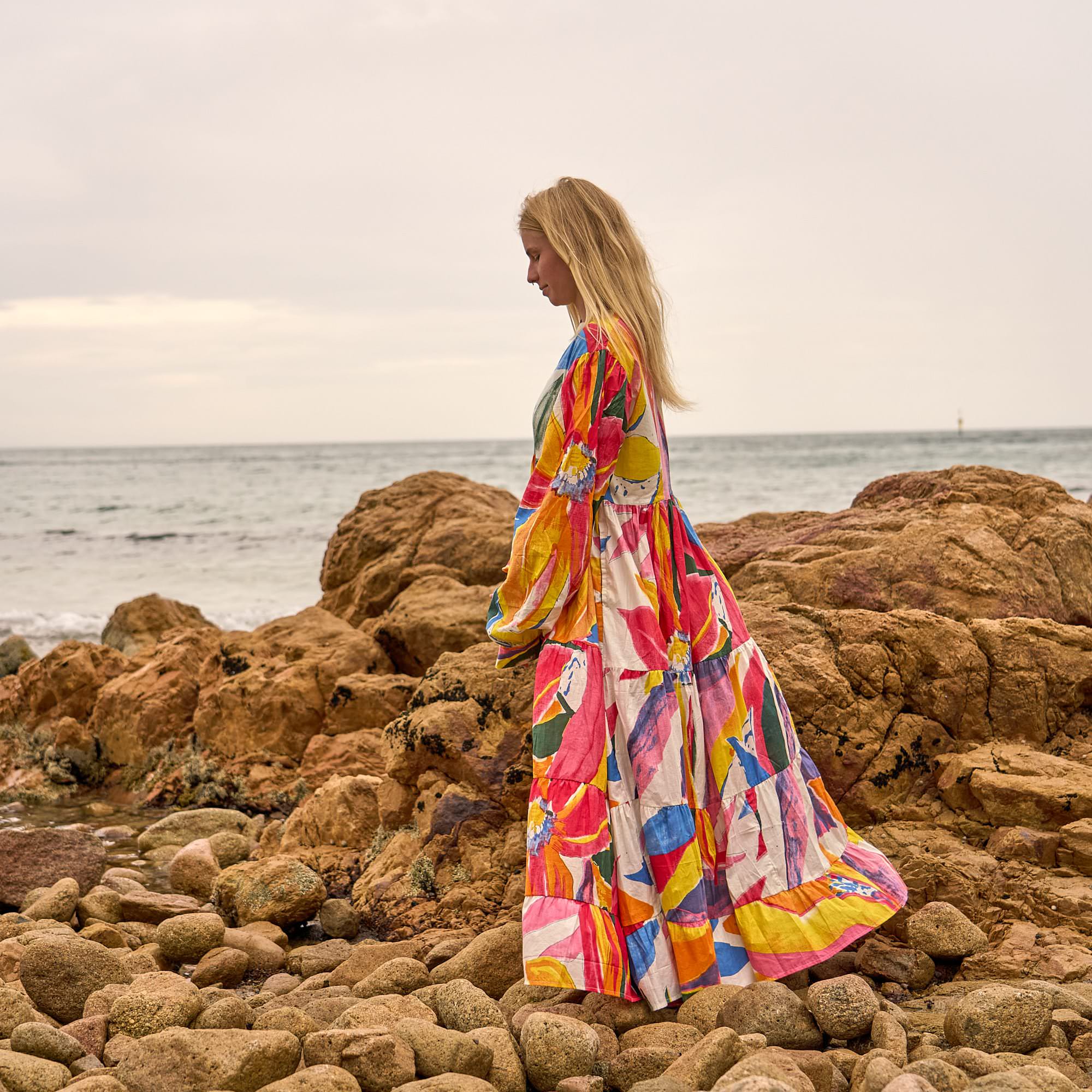 Woman wearing a colorful abstract print cotton maxi dress with balloon sleeves, standing barefoot on rocky beach with wind-blown hair