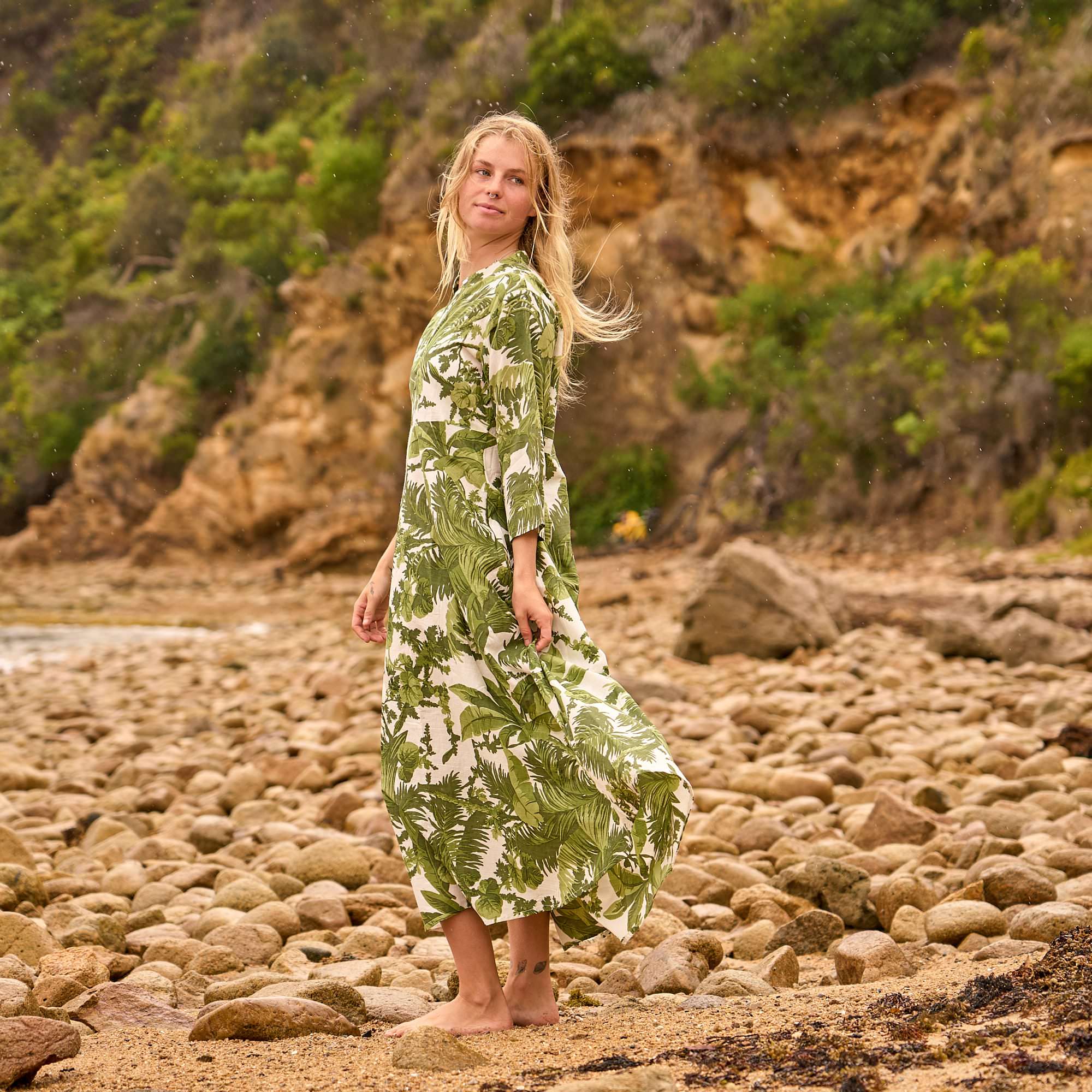 Blonde woman wearing a hand-printed green tropical cotton dress, walking barefoot on the beach in a relaxed maxi silhouette