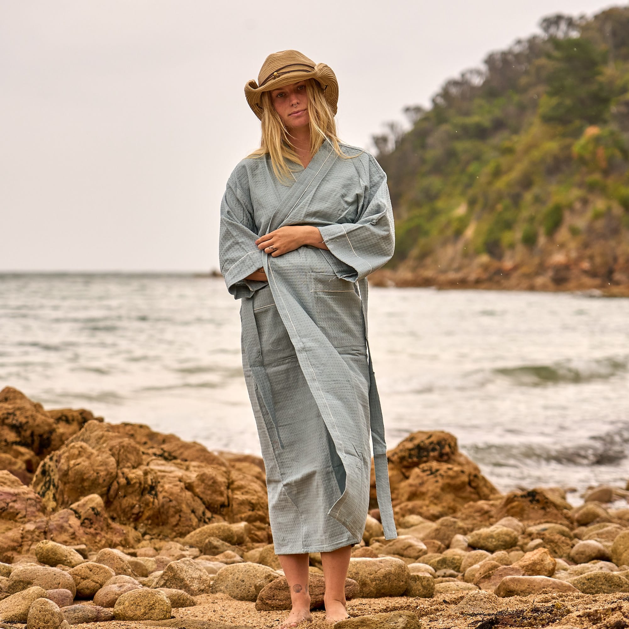 Woman wearing a sage blue waffle cotton robe and straw hat, standing barefoot on a rocky beach with ocean waves in the background.