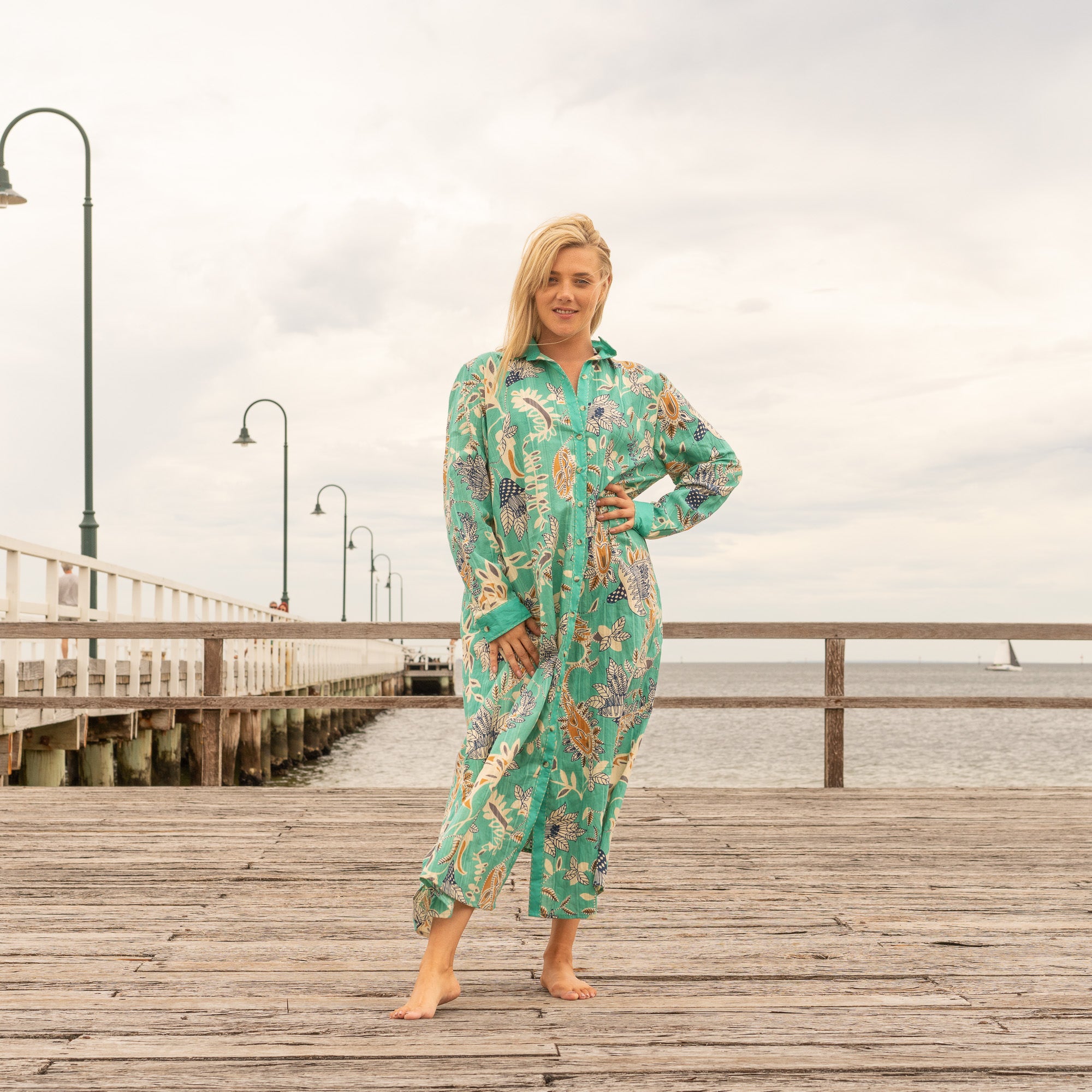 Woman wearing a green botanical print cotton maxi shirt dress, barefoot on a wooden pier with the ocean in the background.