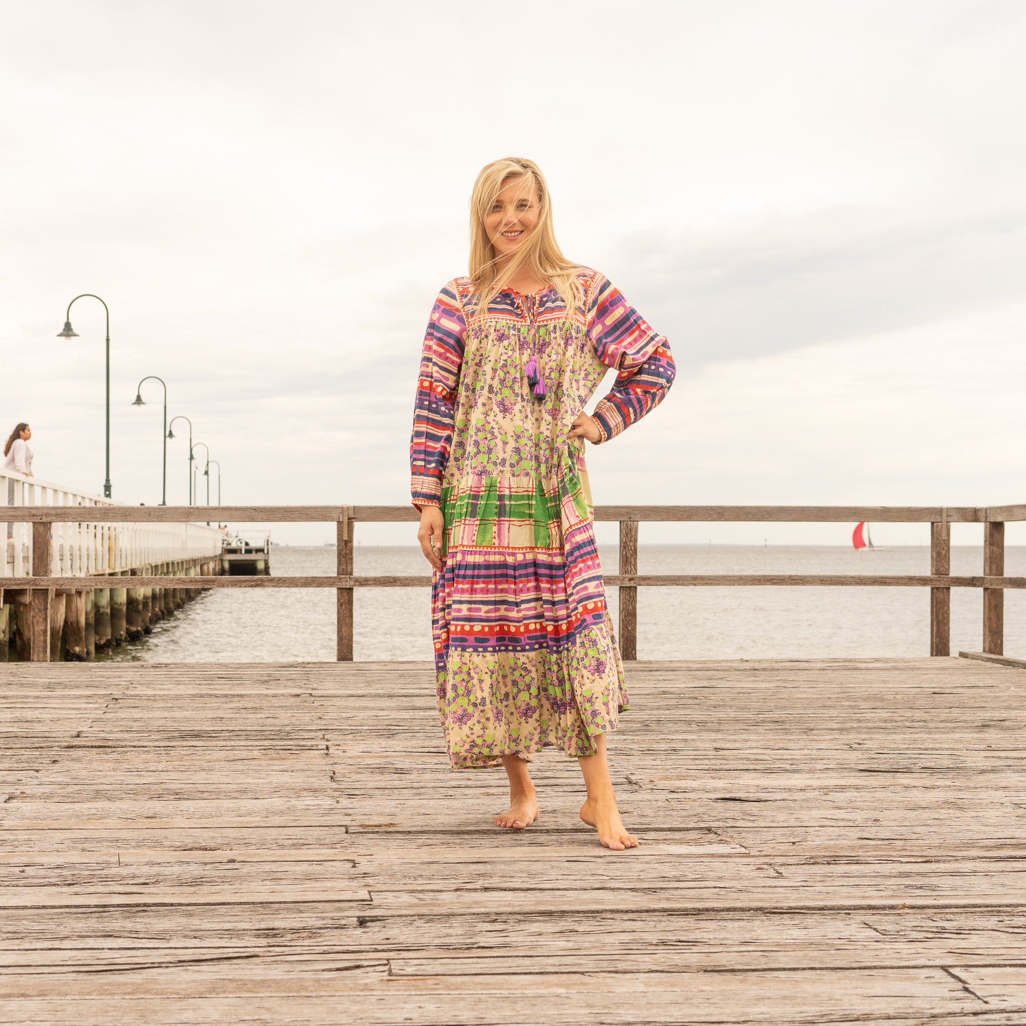 Blonde woman wearing a colorful boho maxi dress on a wooden boardwalk by the sea, smiling with the wind in her hair.