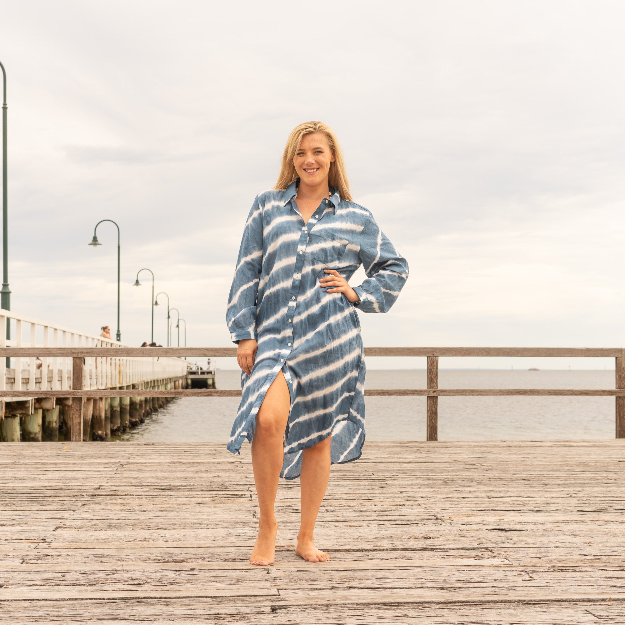 Smiling woman wearing a long-sleeve indigo blue hand-dyed cotton shirt dress, walking barefoot on a wooden pier with the sea in the background.