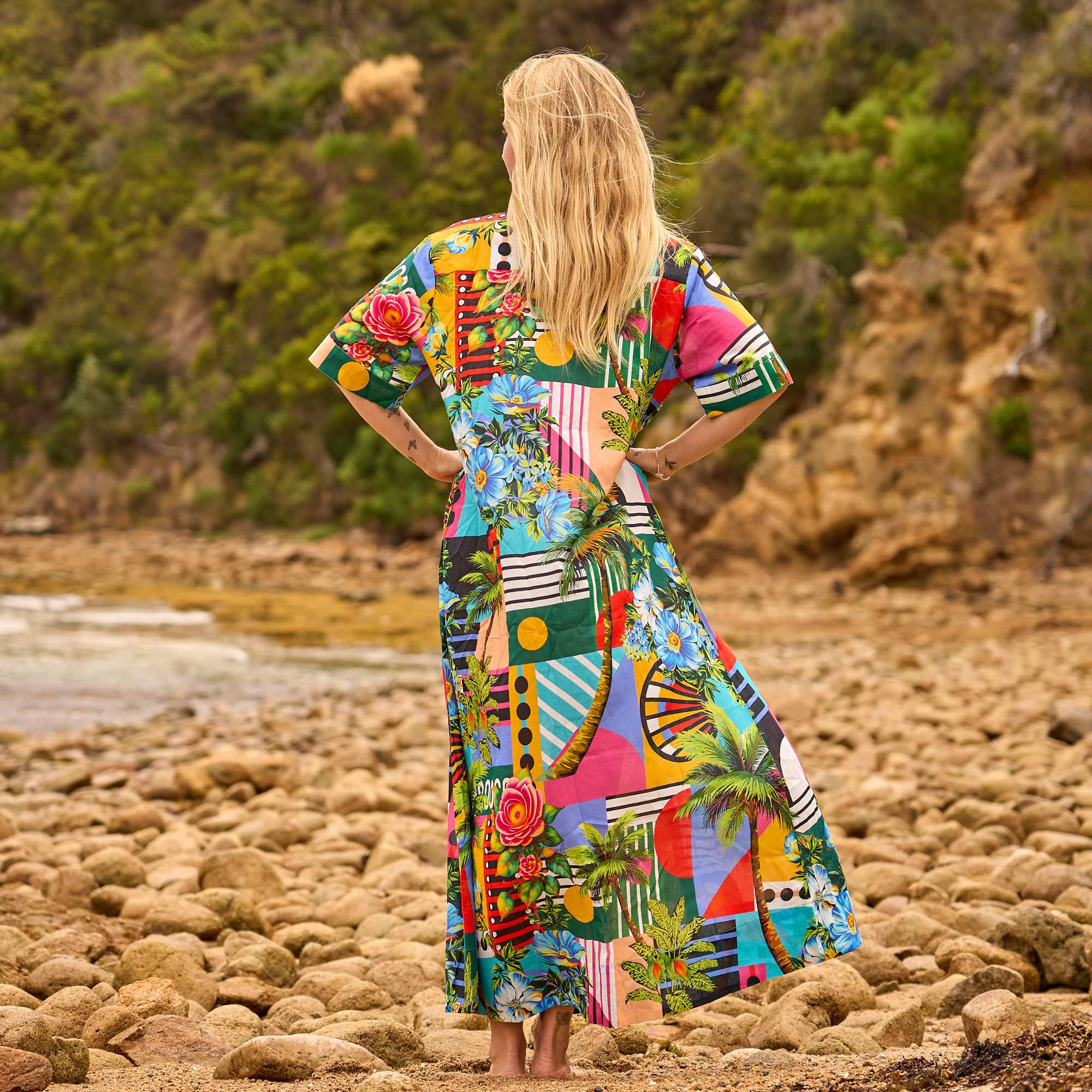 Woman in a colorful dress standing on a rocky beach with greenery in the background