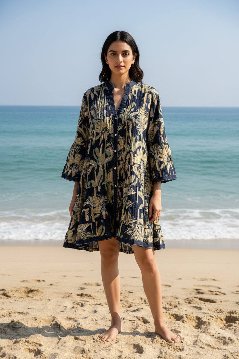 Model wearing a navy and olive hand-printed tropical mini cotton dress with tiered hem and full front buttons.