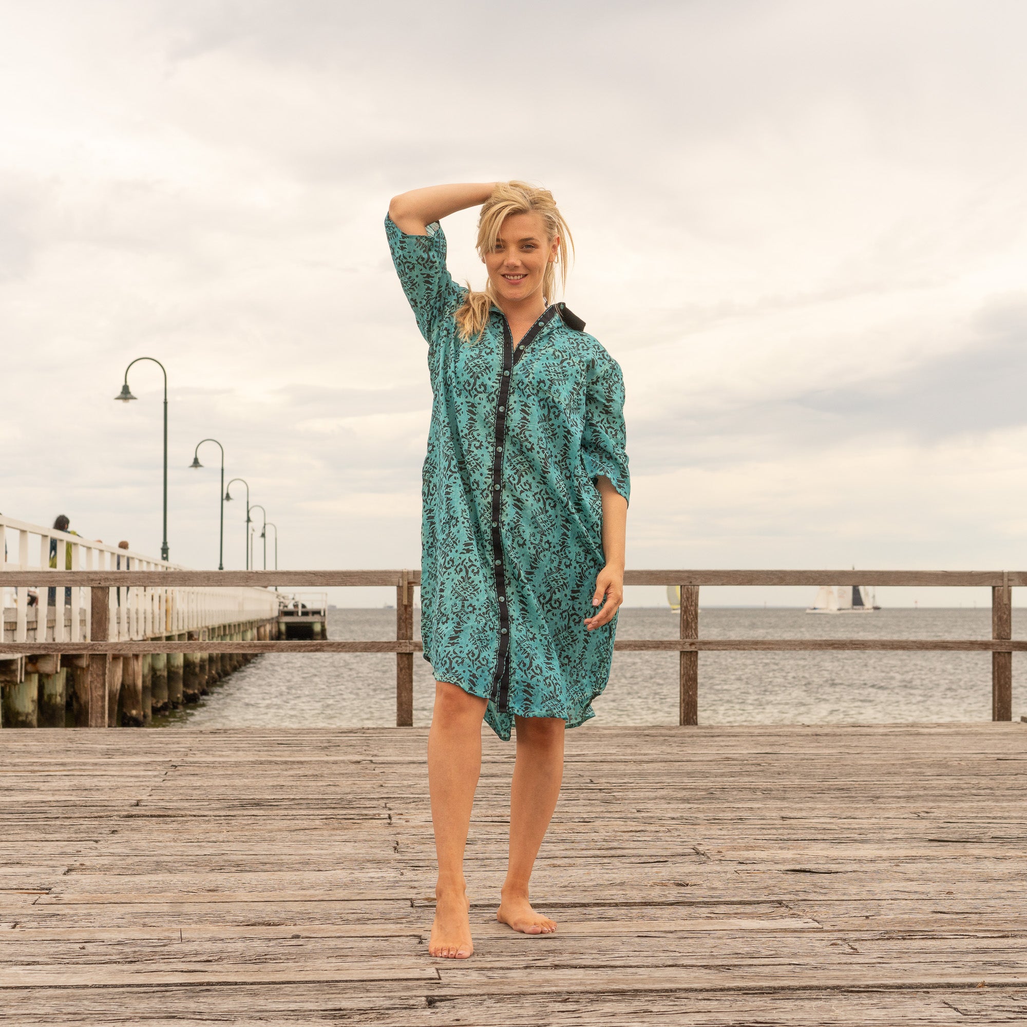 Woman in a teal printed cotton shirt dress with black trim, walking barefoot on a wooden pier by the sea on a cloudy day.