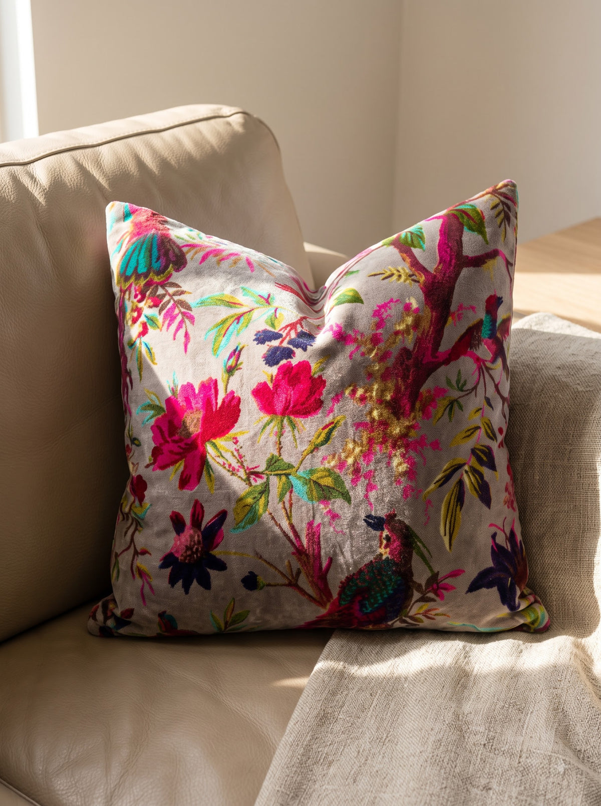 A square throw pillow with a vibrant, multicolored embroidered floral and bird pattern on a beige linen background, styled on a beige leather sofa in warm natural light. Floral bird velvet cushion cover styled on tan leather sofa
