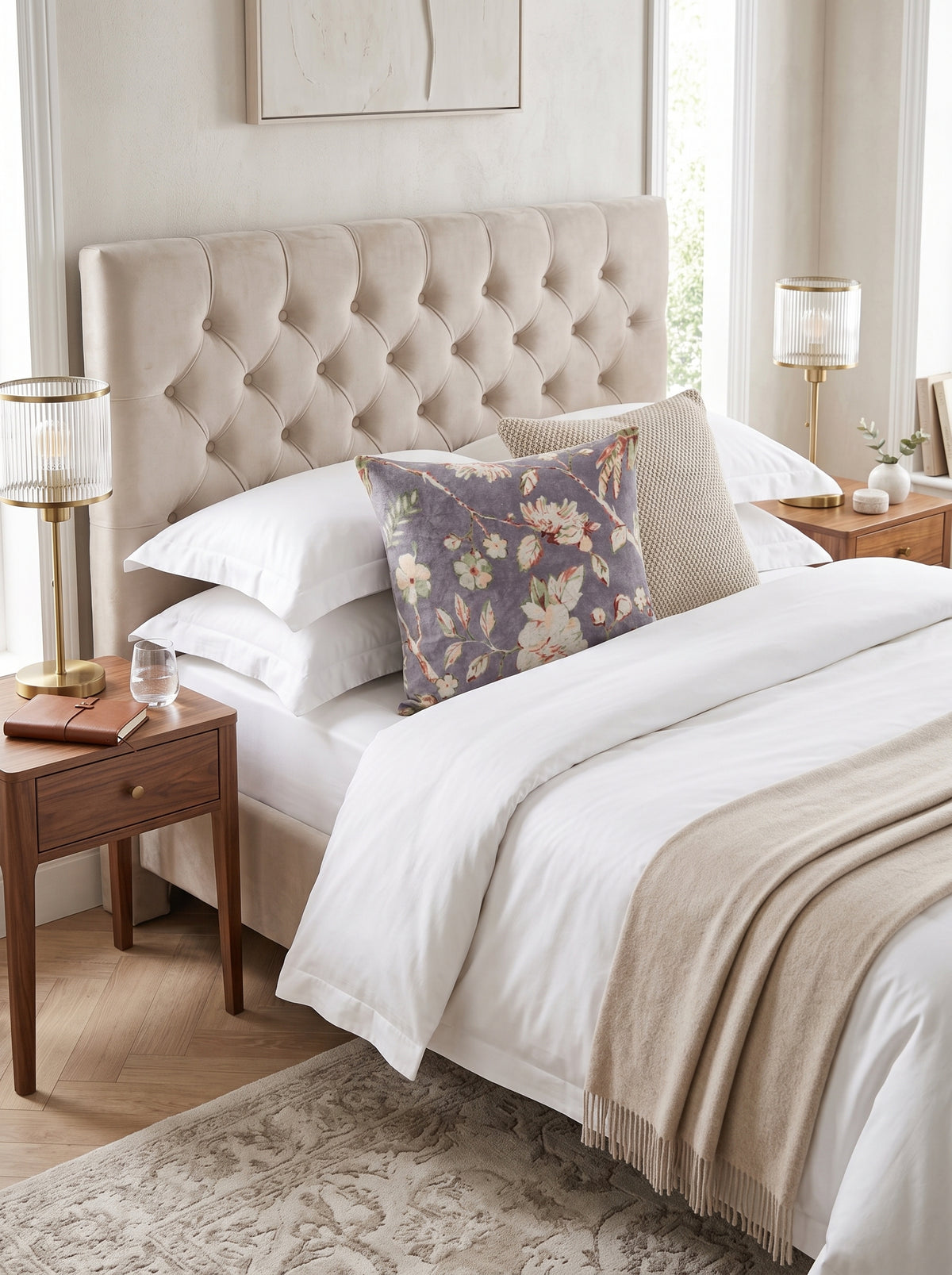 A styled bedroom scene featuring a bed with a tufted beige upholstered headboard, white bedding, a beige throw blanket, and a decorative floral accent pillow. The bed is flanked by matching wooden nightstands with gold table lamps, set against a warm neutral wall.
